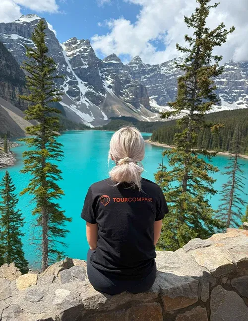 Een vrouw in een TourCompass-T-shirt kijkt uit over een turquoise meer en bergen met besneeuwde toppen in Canada
