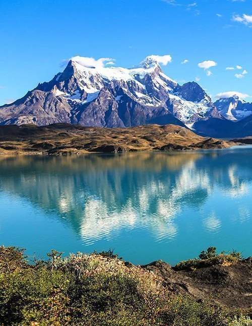 Een spiegelglad bergmeer, waarin sneeuwbedekte toppen weerspiegelen in het turquoise water onder een wolkeloze hemel.